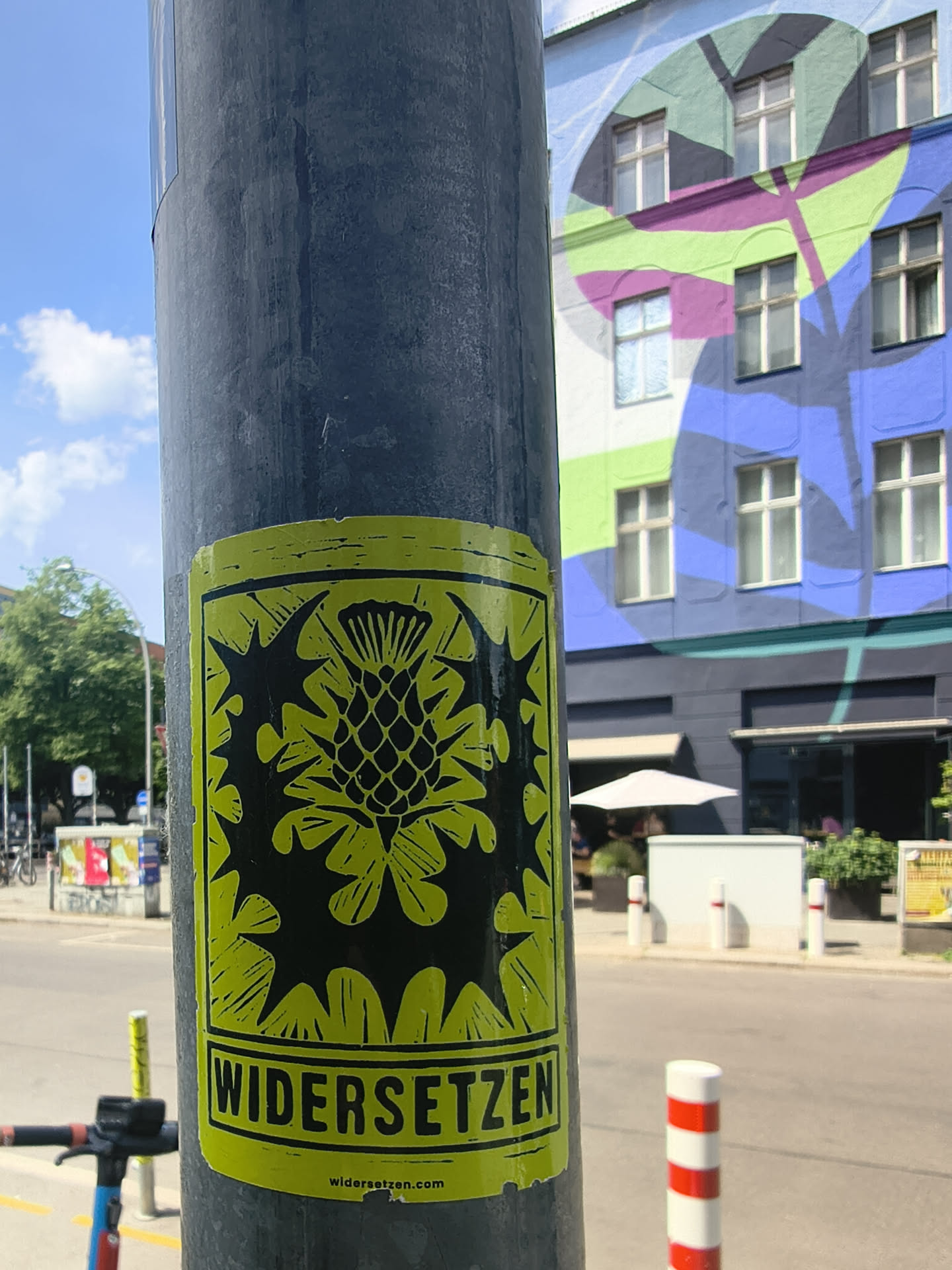 A rectangular lime-green sticker on a pole. It displays a black, linocut-style graphic of a thistle. The bottom features the bold text 'WIDERSETZEN' and the website 'widersetzen.com' in small print.