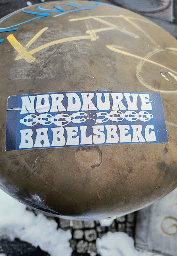 A sticker featuring the text "Nordkurve Babelsberg" in a retro font, with a graphic of a broken chain underneath. The sticker is affixed to a weathered surface.