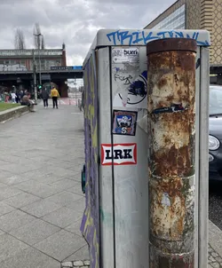 Street sticker The sticker at the center of the image features two main elements. The top element is a square sticker with a stylized blue and white logo, featuring abstract shapes and text that is mostly illegible but appears to say 'BURRI'. Below this is a rectangular sticker with a red border and a white background, containing the bold black text 'LRK'. Adjacent to this is another square sticker depicting a cartoonish character in blue and white, with a retro aesthetic and the text 'FANATICS' and 'GST 2002' visible.