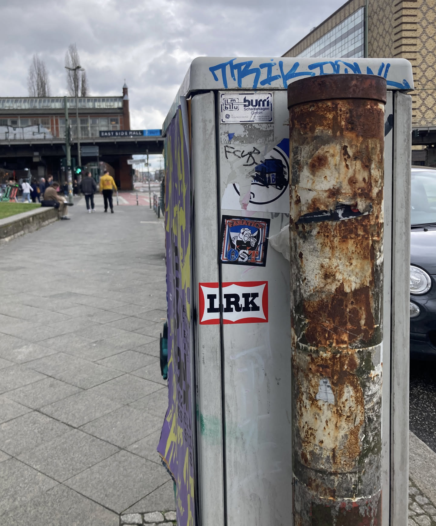 The sticker at the center of the image features two main elements. The top element is a square sticker with a stylized blue and white logo, featuring abstract shapes and text that is mostly illegible but appears to say 'BURRI'. Below this is a rectangular sticker with a red border and a white background, containing the bold black text 'LRK'. Adjacent to this is another square sticker depicting a cartoonish character in blue and white, with a retro aesthetic and the text 'FANATICS' and 'GST 2002' visible.