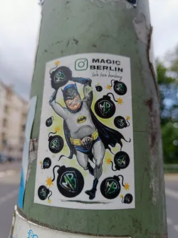 Street sticker A sticker featuring a stylized illustration of Batman defusing bombs.  The design includes the text "MAGIC BERLIN" and "We love bombing", suggesting a playful, artistic commentary. Several bombs feature a stylized "MB" logo. An Instagram icon is also present. 