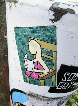 Street sticker The sticker features an illustration of a person seated in a chair, engrossed in a book with the provocative title 'The Clit'. The figure is simply drawn with few details, set against a textured background. Other stickers are partially visible on the post, including one with the text 'SON GOKU'.