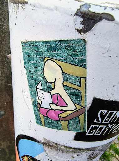 The sticker features an illustration of a person seated in a chair, engrossed in a book with the provocative title 'The Clit'. The figure is simply drawn with few details, set against a textured background. Other stickers are partially visible on the post, including one with the text 'SON GOKU'.