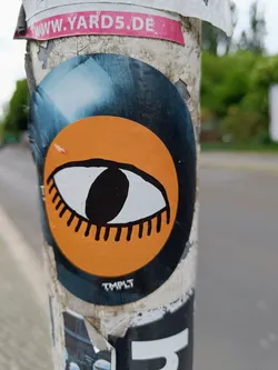 Close-up of a sticker featuring a stylized eye graphic on an orange circle. The sticker is adhered to a weathered pole, and other stickers are visible around it.  The artist's mark 'TMPLT' is barely visible at the bottom. 