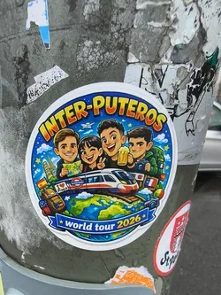 A circular sticker with a white border, featuring a colorful, cartoonish illustration of four friends on a 'world tour'. The central image depicts a high-speed train traveling across a globe, surrounded by various European landmarks such as the Eiffel Tower and the Leaning Tower of Pisa. Four smiling characters are positioned above the train: one holding a map, another winking, a third holding a mug of beer, and the fourth with a backpack and camera. The background is a starry blue sky with clouds. Small flags (Spain, France) and other travel items like luggage are scattered around the globe. The top of the sticker prominently displays 'INTER-PUTEROS' in orange and yellow bubble letters, while a blue banner at the bottom reads 'world tour 2026' flanked by yellow stars. The overall aesthetic is cheerful and adventurous.
