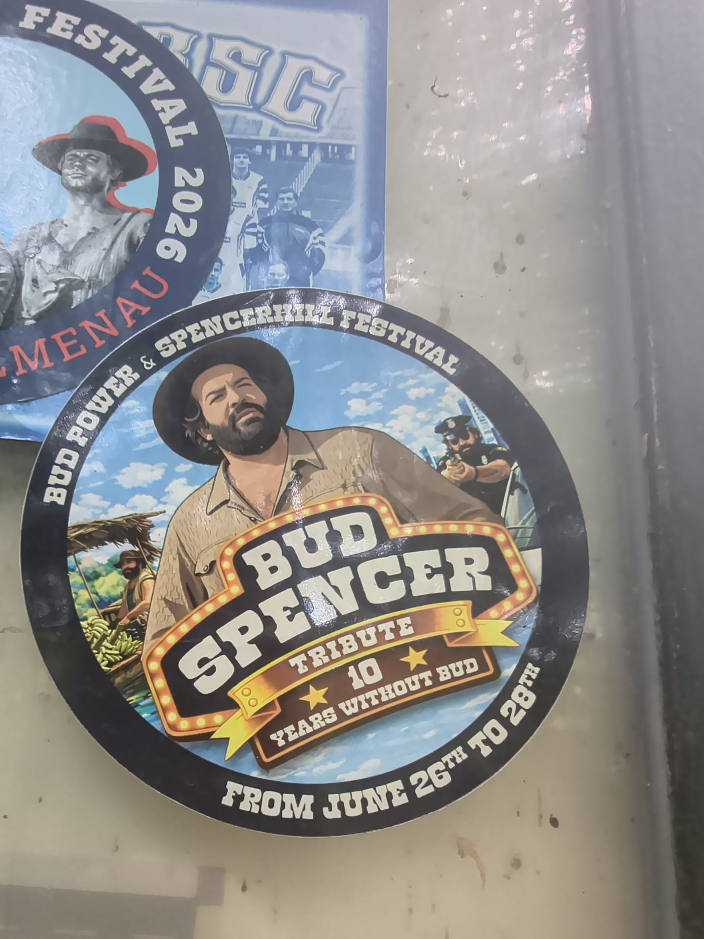 Circular sticker featuring an illustrated portrait of Bud Spencer. Includes a marquee-style tribute sign, a police officer, and a tropical background under a blue sky.