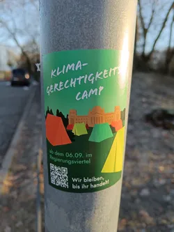 Street sticker A sticker promoting a climate justice camp starting on September 6th in the government district.  The sticker features an illustration of tents pitched in front of a government building. A QR code is included.