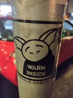 Street sticker A sticker featuring a simple cartoon character with small horns or ears, a round face, and a slightly mischievous expression. The text "WARM INSIDE" is prominently displayed below the character. The sticker also includes the text "MUGGLE-BERLIN" at the bottom. The character appears friendly and somewhat whimsical.