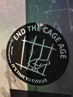 Street sticker A round black sticker with white text and a drawing of a bird in a cage. The text says "End the Cage Age" at the top and "It's time to evolve" at the bottom. There is also a website address and a place to sign.