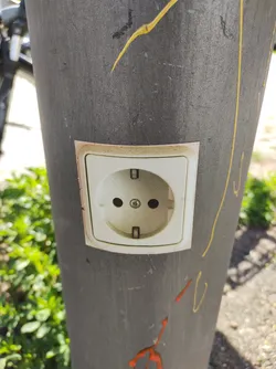 This sticker depicts a realistic image of a European-style electrical outlet. It is affixed to a gray pole, adding a humorous and unexpected element to the urban environment. The sticker's simple yet effective design stands out against the pole's backdrop. 