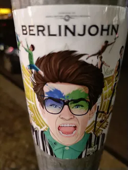 Street sticker A colorful sticker featuring an illustration of a person with paint splatters on their face and glasses, with a bold expression. The background includes various figures in motion, adding a dynamic feel. The name "BERLINJOHN" is prominently displayed at the top. 