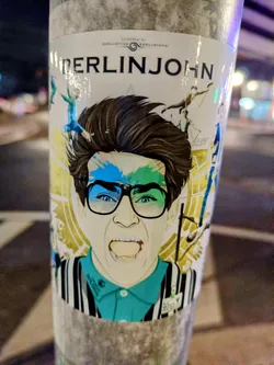 Street sticker A sticker featuring an illustration of Berlinjohn, with a vibrant design and detailed artwork. The central figure is a person with glasses, expressive features, and stylized hair. Surrounding elements add depth and artistic flair.  The sticker is affixed to a pole.