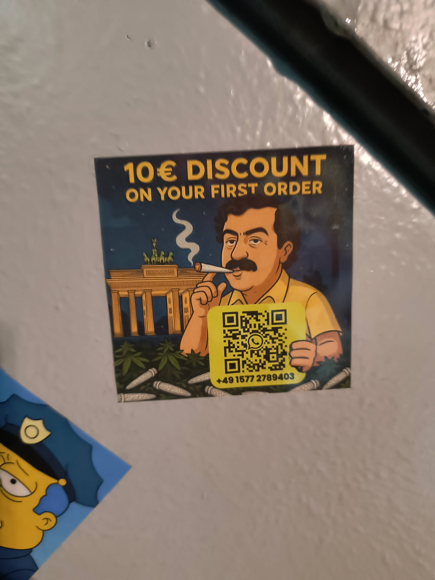 Street sticker The sticker features a cartoon man with a dark mustache and hair, wearing a yellow shirt, smoking a joint. He holds a prominent yellow square QR code. In the background, the Brandenburg Gate in Berlin is depicted under a dark blue night sky, with green cannabis plants and several white pre-rolled joints scattered in the foreground. The top of the sticker displays bold yellow text, and a phone number is printed below the QR code. The overall color palette includes dark blue, yellow, green, and brown.