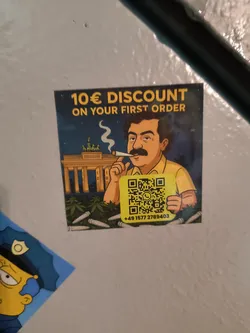 Street sticker The sticker features a cartoon man with a dark mustache and hair, wearing a yellow shirt, smoking a joint. He holds a prominent yellow square QR code. In the background, the Brandenburg Gate in Berlin is depicted under a dark blue night sky, with green cannabis plants and several white pre-rolled joints scattered in the foreground. The top of the sticker displays bold yellow text, and a phone number is printed below the QR code. The overall color palette includes dark blue, yellow, green, and brown.