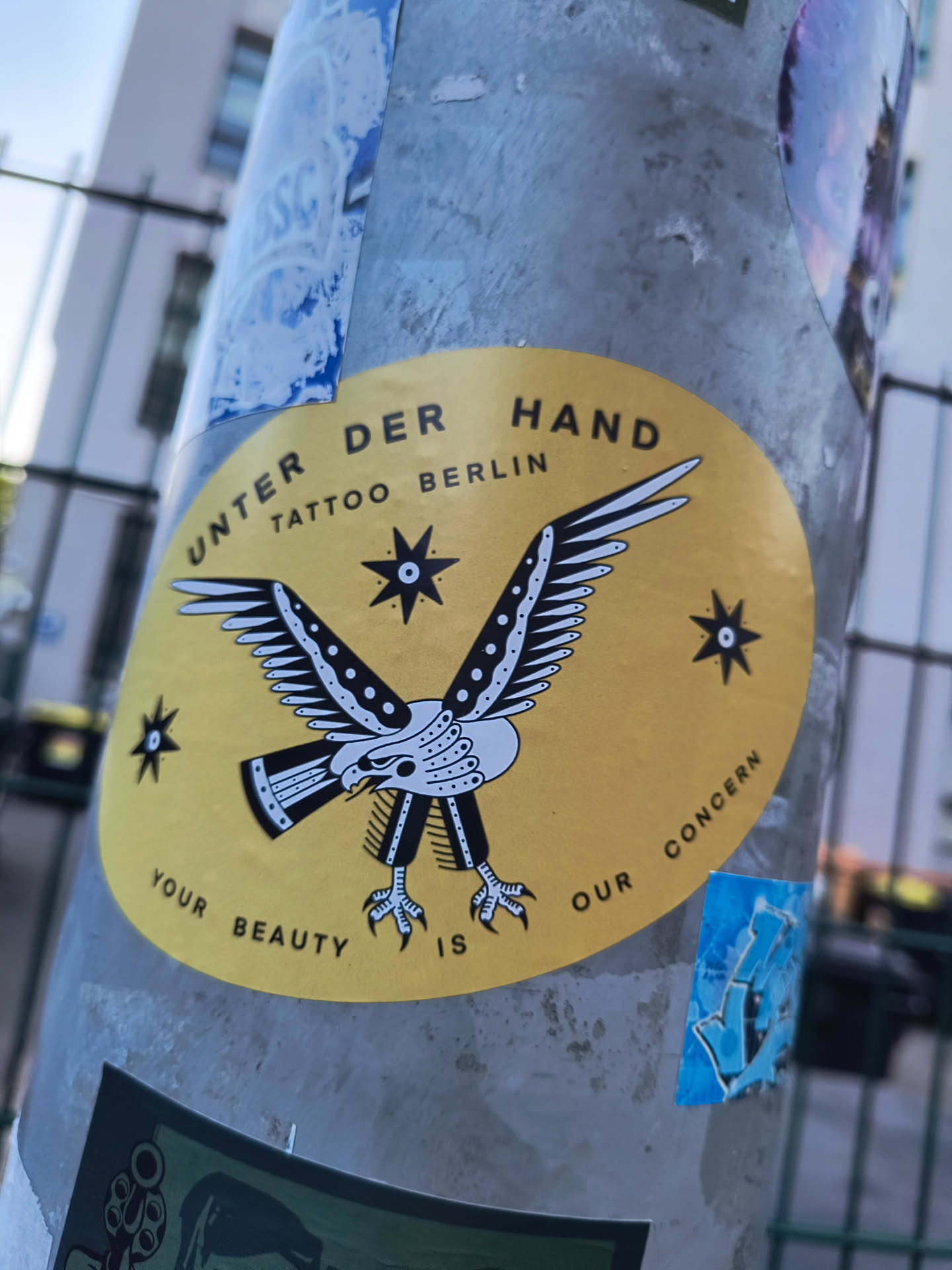 This sticker, attached to a pole, has a vintage vibe. The background is a muted yellow, and the main graphic is a black and white eagle with its wings spread. The eagle is stylized, with intricate details on its feathers and legs. Above the eagle, the sticker reads 'UNTER DER HAND TATTOO BERLIN.' Below the eagle, it reads 'YOUR BEAUTY IS OUR CONCERN.'