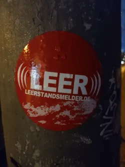 A red circular sticker is affixed to a gray pole. The sticker displays the word "LEER" prominently in white uppercase letters, with stylized sound wave symbols on either side.  Below "LEER" is the smaller text "leerstandsmelder.de". The sticker appears slightly damp, showing water droplets on its surface.