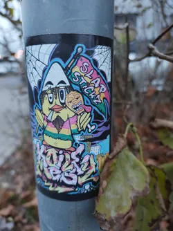 Street sticker A vibrant sticker featuring a cartoon bird character holding a banner that says 'Slap Sticky'. The character has a unique design, with a large head and beak, and is surrounded by a web-like design. The style is reminiscent of 90s cartoons and graffiti art.  The text 'KOIS KRISY' is visible in graffiti-style lettering at the bottom. The overall color palette is bright and playful. 
