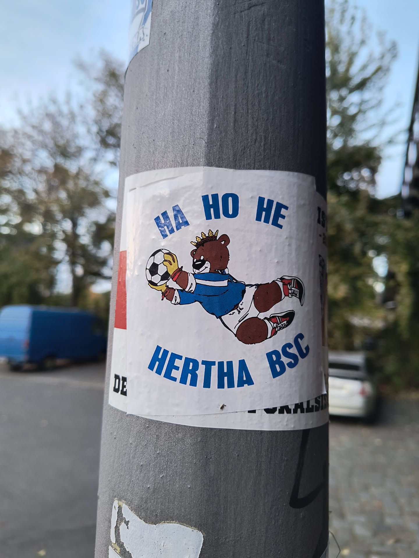 The sticker features a cartoon bear wearing a blue and white football jersey, white shorts, and red and white sneakers. The bear has a crown on its head and is depicted in a dynamic pose, catching a black and white soccer ball. The text 'HA HO HE' is written in blue capital letters above the bear, and 'HERTHA BSC' is written in blue capital letters below it. The sticker has a white background with some visible texture and slight creasing, indicating it is indeed a sticker applied to a surface.