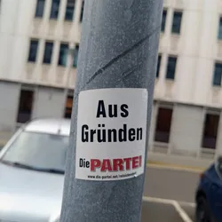 Street sticker A sticker featuring the text "Aus Gründen" (For Reasons) and the logo of the German satirical party "Die PARTEI". The sticker is affixed to a gray pole.