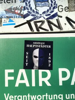Street sticker The sticker shows the text "ASOZIALE HAUPTSTÄDTER SEIT 1892" above an image of a mask divided vertically, one half white and the other dark blue. The text translates to "Antisocial Capital Citizens Since 1892."