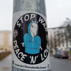 A round sticker with a unicorn wearing a blue hoodie, with the text 'STOP WAR PEACE 'N' LOVE' surrounding it. The sticker also includes peace symbols and is attributed to 'Street Art Thessaloniki, Greece'.