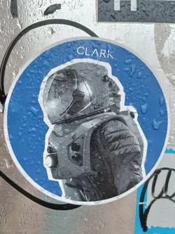 Street sticker Close-up of a round sticker featuring a grayscale astronaut illustration. The astronaut is wearing a helmet and spacesuit with visible details. The sticker has a blue circular background with water droplets visible on its surface. The word 'CLARK' is printed at the top of the sticker.