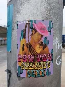Street sticker A sticker featuring a man in a pink cowboy hat, holding a whip and a gun. The background is purple with cacti. The text 'Pow Pow Sheriff Sexy' is prominently displayed.