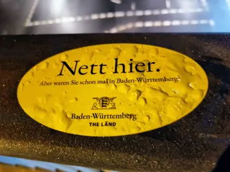 Yellow oval sticker with the text "Nett hier." (Nice here.) and  "Aber waren Sie schon mal in Baden-Württemberg?" (But have you ever been to Baden-Württemberg?). It also shows the coat of arms of Baden-Württemberg and the text "Baden-Württemberg THE LÄND". The sticker seems to be attached to a dark surface and has water droplets on it.