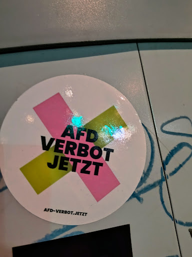 Street sticker A round sticker with a pink and yellow X design, overlaid with the words "AFD VERBOT JETZT" in bold black letters. The website "AFD-VERBOT.JETZT" is printed at the bottom of the sticker.