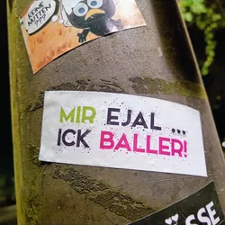 Street sticker A white sticker with the text "Mir egal... ick baller!" in different colors. The text is written in a playful, slightly rebellious style.  The sticker is affixed to a textured, dark-greenish grey surface, likely a pole or similar.