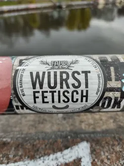 Street sticker Round sticker with the text "Wurst Fetisch" in the center. It also contains the logo of Fabis Esskultur and some additional text in a smaller font. The sticker is placed on a pole.