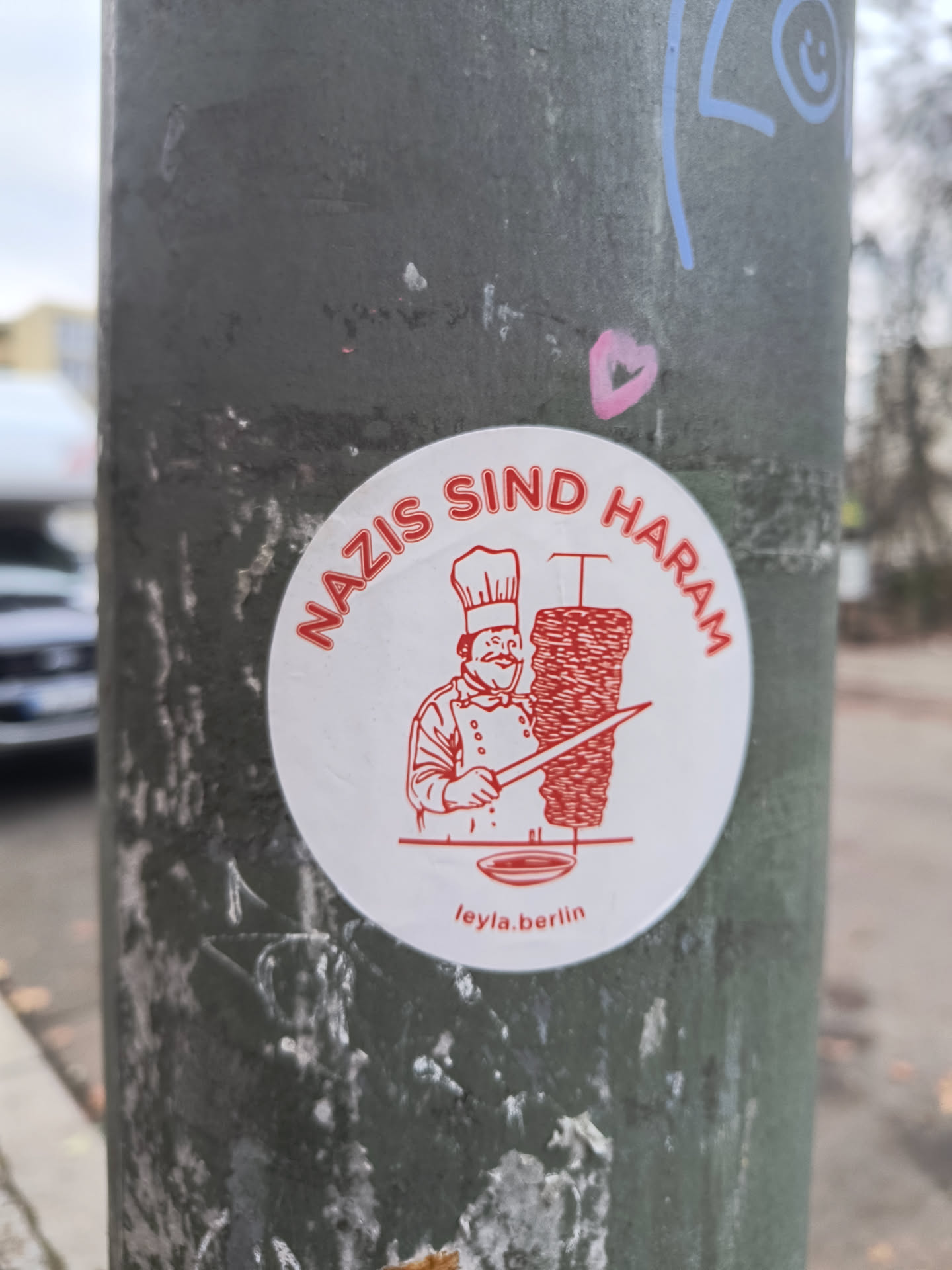 Street sticker A circular white sticker featuring a red line-art illustration. The central image depicts a smiling, portly chef wearing a tall chef's hat and a double-breasted jacket, holding a large knife. To the chef's right is a tall, vertical spit of döner kebab meat, with a small drip tray beneath it. Arcing around the top edge of the circle, in red capital letters, is the German phrase "NAZIS SIND HARAM". At the bottom, in smaller red text, is "leyla.berlin". The overall style is cartoonish and bold, with a clear, direct message.