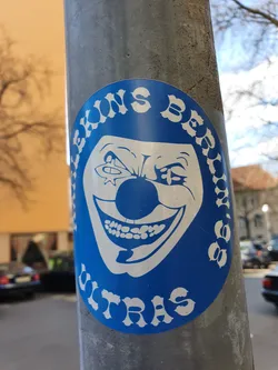 Street sticker A blue circular sticker featuring a grinning clown face with unsettling details. The text around the image seems to read "LEBONS BERLIN 999 UTRAS". The style is reminiscent of vintage or retro designs, creating a unique and slightly disturbing aesthetic. The sticker is affixed to a metal pole in an urban environment.