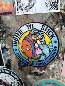 Street sticker This round sticker shows a cartoon fish smoking and a light blue monster in the middle. The text says "UNITED WE STICK * GREETINGS FROM COLOGNE *" around the edge of the sticker.