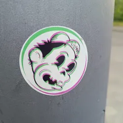 A round sticker featuring a cartoon skull character with a playful expression. The skull is designed in a glitch art style with pink and green outlines. The sticker is stuck on a gray pole.