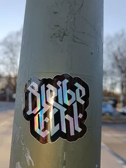 A black sticker with a holographic effect features the text "Bleib echt" in a stylized Gothic font. The sticker is affixed to a weathered green pole, suggesting it's been placed outdoors. The background is slightly blurred, showing a street scene with trees and vehicles.