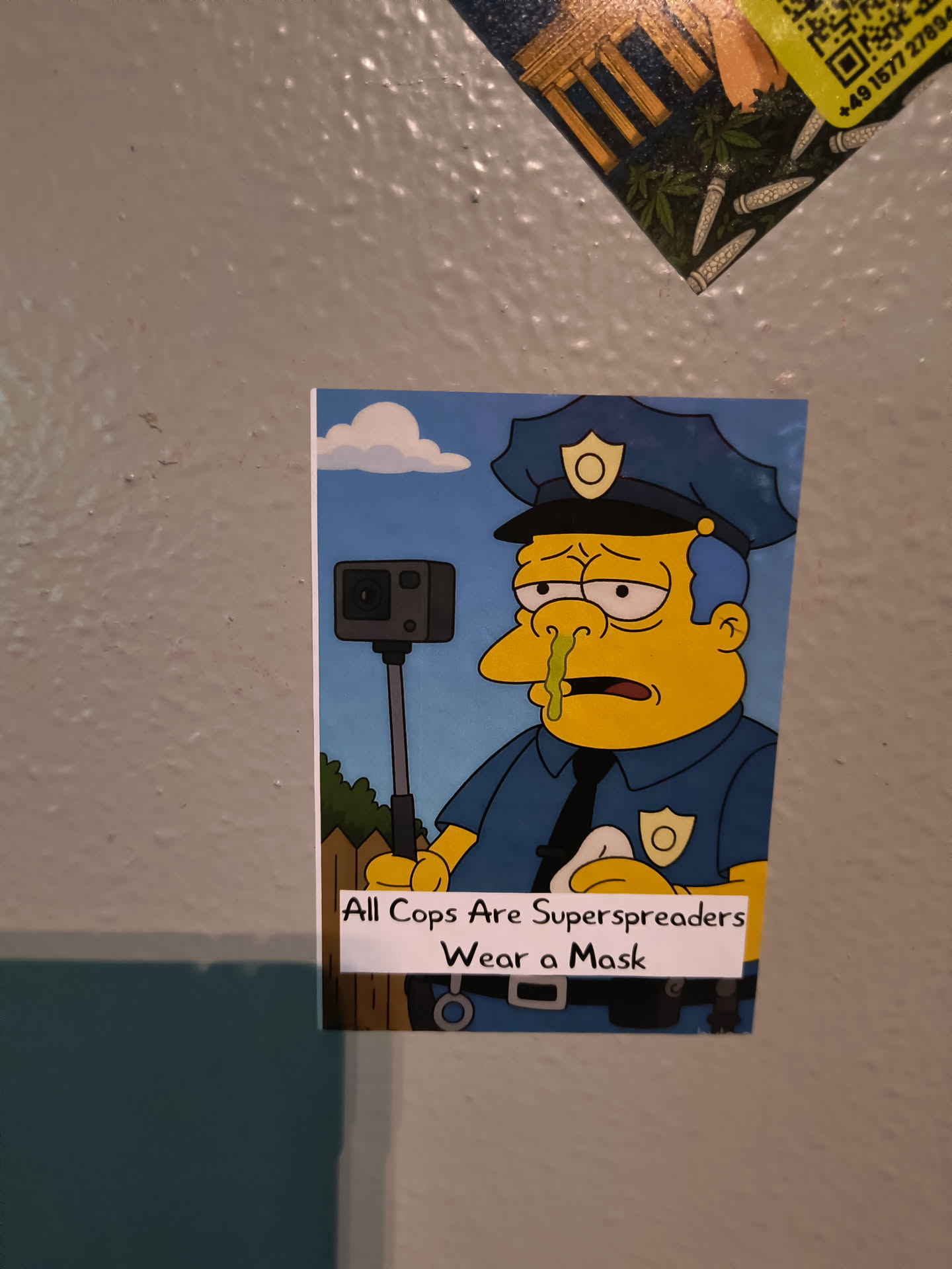 Street sticker The sticker features a cartoon illustration of Chief Wiggum from The Simpsons, depicted as a police officer in a blue uniform, complete with a hat and badge. His face is yellow, and he has a prominent runny green nose, suggesting illness. He holds a black GoPro camera on a stick in his left hand. The background is a simple blue sky with white clouds and a brown wooden fence. A white banner at the bottom contains black text.