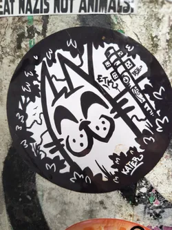 Street sticker The sticker is circular and features a high-contrast black and white illustration. At its center is a stylized, smiling cat's face with large eyes and a wide, upturned mouth, reminiscent of a cartoon character. The cat's body is partially visible, blending into the surrounding elements. The background is a mix of organic, wavy shapes representing foliage and trees on the left and bottom, and more geometric, tower-like structures with small windows or blocks on the right side. Small 'W' shapes are integrated into the tree lines. The overall style is playful and somewhat abstract, with clean lines forming the white elements against a solid black background.