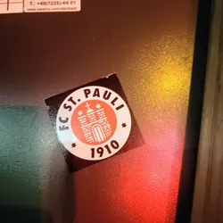 Street sticker A round sticker featuring the logo of FC St. Pauli, a German football club. The logo depicts a stylized gate with three towers and a cross on top, set within a circular border with the club's name and founding year (1910). The sticker is dark-colored with the logo in shades of orange and white.