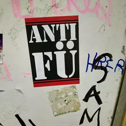 Street sticker A black and red sticker with the text "ANTI FU" in white stencil lettering. The sticker is affixed to a wall with other graffiti and stickers.