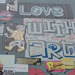 A sticker featuring a bear in a gold design. The sticker is part of a larger collection of stickers on a wall, some with political messages.  The word 'Love' with a musical note and heart, 'with' and 'Art' with scissors and a paint palette are visible on the wall around the bear sticker.