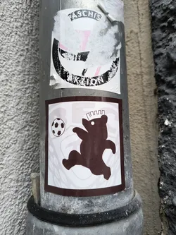 Street sticker A sticker featuring a playful illustration of a bear kicking a soccer ball. The bear is depicted in a dynamic pose, suggesting movement and action. The sticker has a simple, minimalist design with a color scheme that includes shades of brown and white.