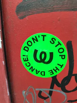 Street sticker A circular, bright neon green sticker with bold black text and a central symbol. The text "DON'T STOP" is arched around the top circumference, and "THE DANCE!" is arched around the bottom. In the center, there's a large, stylized black symbol resembling a double 'U' or 'W'. The sticker appears slightly worn and is affixed to a textured, reddish-orange surface with black and silver graffiti marks.
