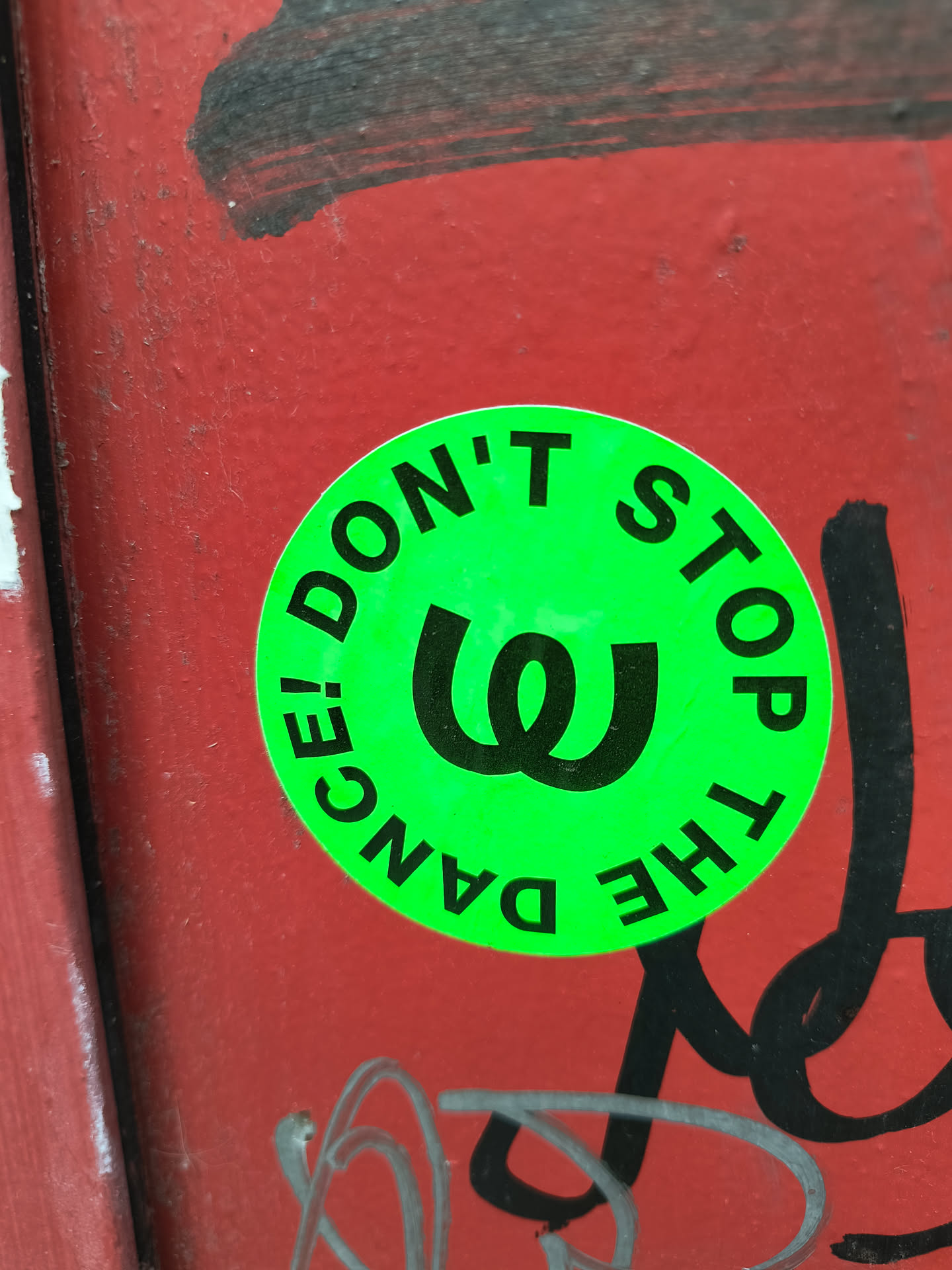 A bright green round sticker with black text that reads "DON'T STOP THE DANCE!" circling around a black letter W like symbol in the middle. The sticker is stuck on a red, graffitied wall.