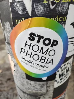 A sticker with a rainbow border and the text "STOP HOMOPHOBIA" in bold black letters. Below it, smaller text reads "Enough is Enough!" with a website address: enough-is-enough.eu. The sticker promotes equality and human rights within the LGBTQIA+ community.