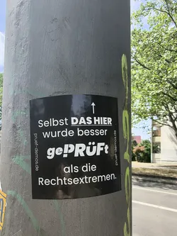 Street sticker A black rectangular sticker affixed to a grey metal pole. It features white German text with an upward-pointing arrow, criticizing right-wing extremists. The URL 'pruef-demos.de' is printed vertically on both the left and right margins.