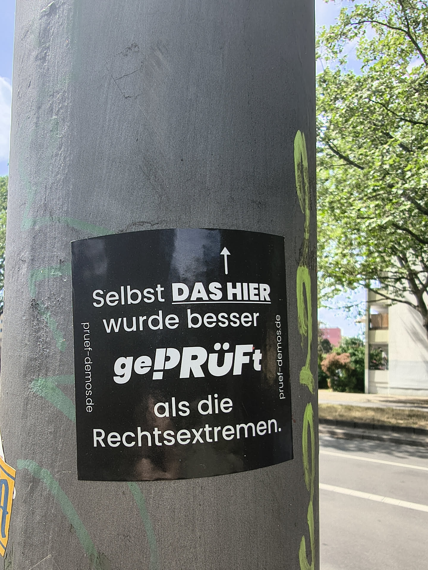 A black rectangular sticker affixed to a grey metal pole. It features white German text with an upward-pointing arrow, criticizing right-wing extremists. The URL 'pruef-demos.de' is printed vertically on both the left and right margins.