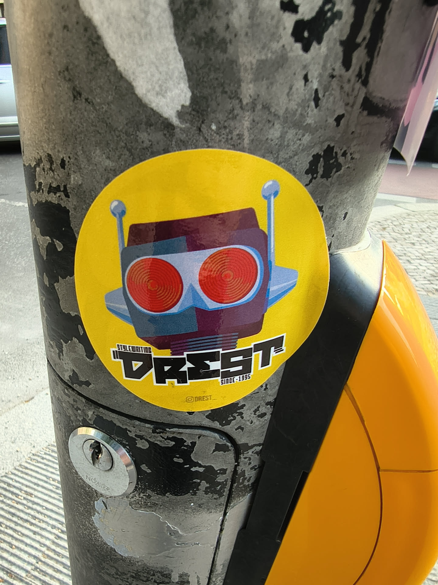 A round yellow sticker depicting a retro robot head with large red eyes and blue antennae. Below the illustration, stylized black-and-white text reads 'DREST', flanked by 'STYLEWRITING' and 'SINCE 1995'. A small Instagram handle is visible at the bottom.