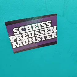 Street sticker A sticker with the text "Scheiss Preussen Munster" on a purple and black background. The text is in white, bold, uppercase letters. The sticker is affixed to a teal wall.