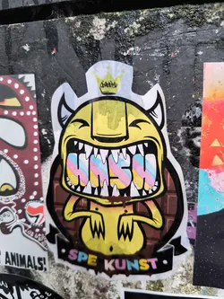 Street sticker The sticker features a stylized, yellow monster-like character with a wide, toothy grin showcasing prominent white fangs. It has small, pointed ears and wears a small, yellow crown with a black scribble design on its head. Inside the monster's open mouth, the word 'HASH' is spelled out in bold, block letters, each letter adorned with a pastel rainbow gradient of pink, blue, and yellow stripes. The character's body is yellow with short, clawed hands. Behind the monster, forming a background element, is a dark brown pattern resembling a chocolate bar, with some brown drips appearing below the mouth area. At the bottom, a black banner or ribbon displays the text 'SPE KUNST' in multi-colored, slightly three-dimensional letters (pink, blue, yellow). The entire design is outlined with a thick black border and a white cut-out edge, indicating it is a die-cut sticker applied to a textured, dark wall.